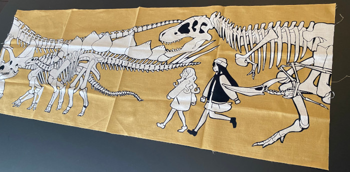 恐竜の骨と2人の女の子のイラストがプリントされたベージュ色生地の手ぬぐいのイメージ画像